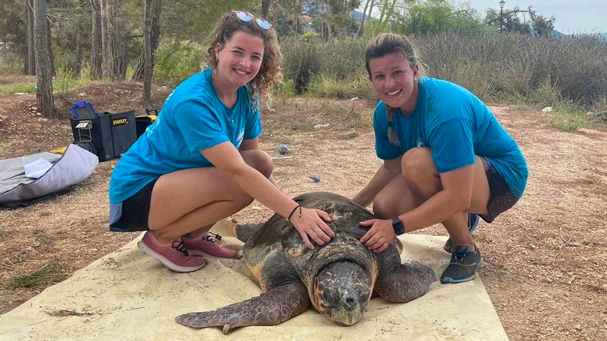 Two women in blue shirts taking a picture with a large sea turtle during a trip to Cabo Verde. Viagens low cost Cabo Verde for eco-friendly travel.